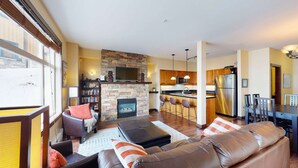 A spacious living room with a stone fireplace, a dining area with a bar, and a kitchen with stainless steel appliances. - Okanagan - The Crescent 104 (Beaverdell)