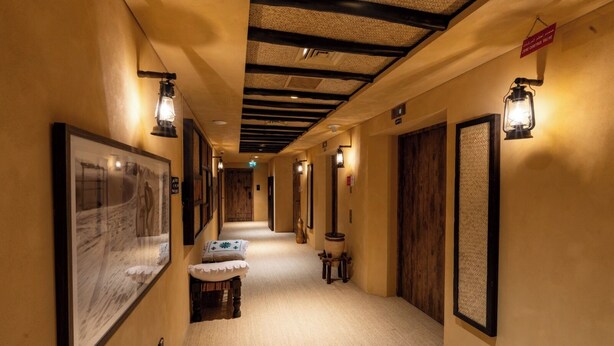A hallway with framed pictures on the walls, a wooden bench, and a sign indicating the room number. - Heritage 2-bedroom Apartment (Dubai)