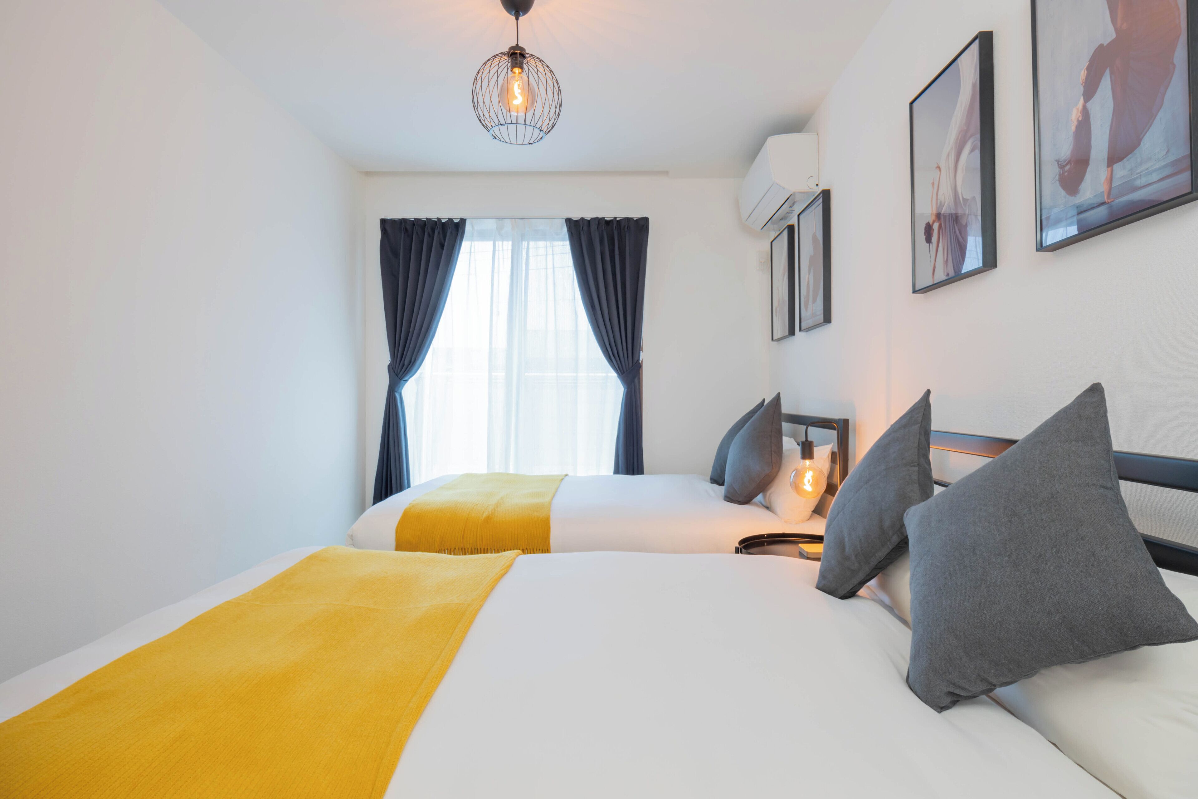 A hotel room with two beds, a yellow throw blanket, grey pillows, a hanging light fixture, and framed artwork on the wall.