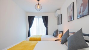 Une chambre d’hôtel avec deux lits, un plaid jaune, des oreillers gris, un luminaire suspendu et des œuvres d’art encadrées accrochées au mur.