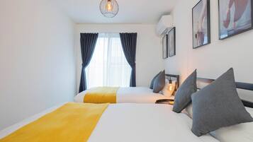 A hotel room with two beds, a yellow throw blanket, grey pillows, a hanging light fixture, and framed artwork on the wall.