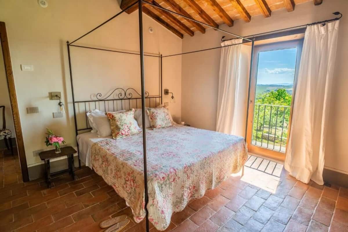 A bedroom with a four-poster bed, a nightstand with a vase of flowers, and a view of greenery through a sliding glass door.
