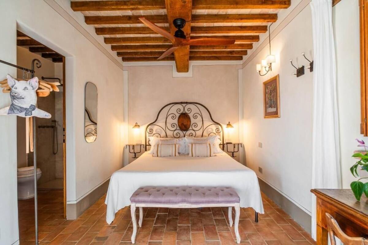 A bedroom with a large bed, wooden ceiling beams, a ceiling fan, and a bench.