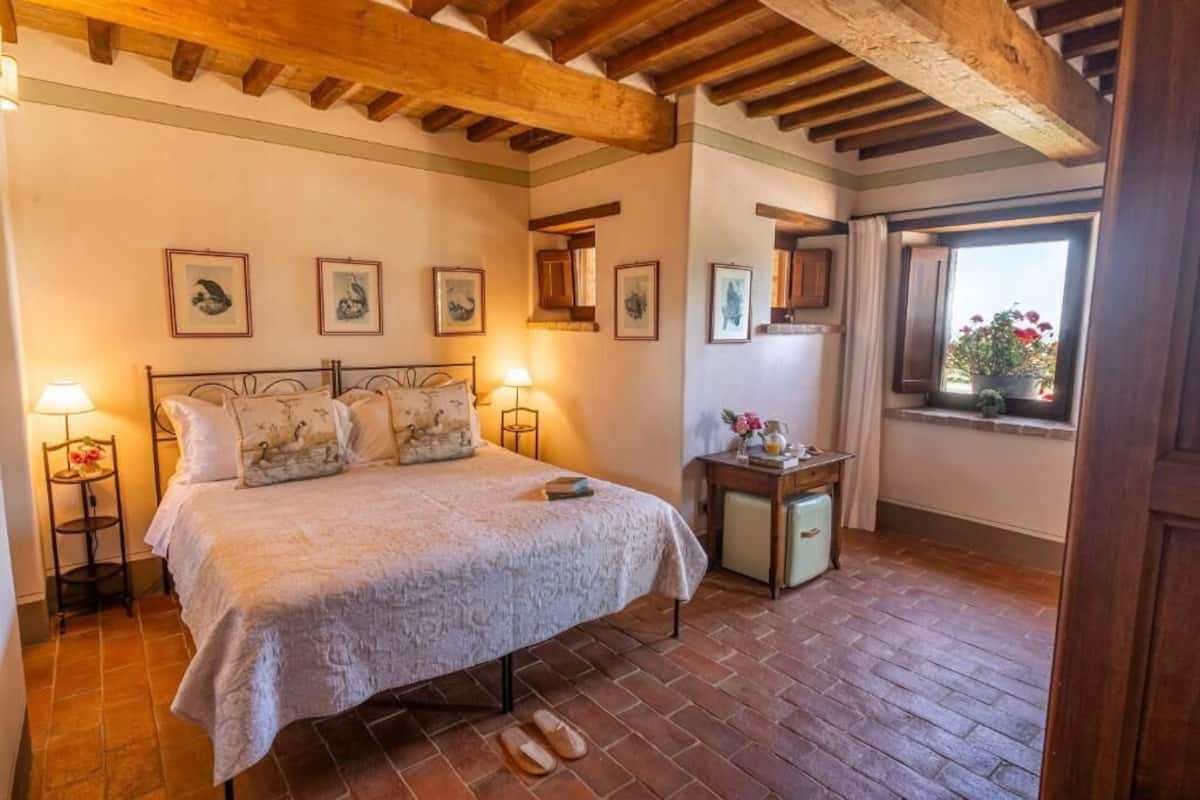 A cozy bedroom with a large bed, wooden beams, a small table with a lamp, and framed pictures on the wall.