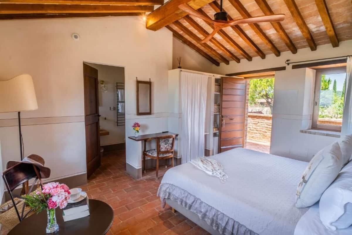 A bedroom with a bed, a desk, a chair, and a table with a vase of flowers.