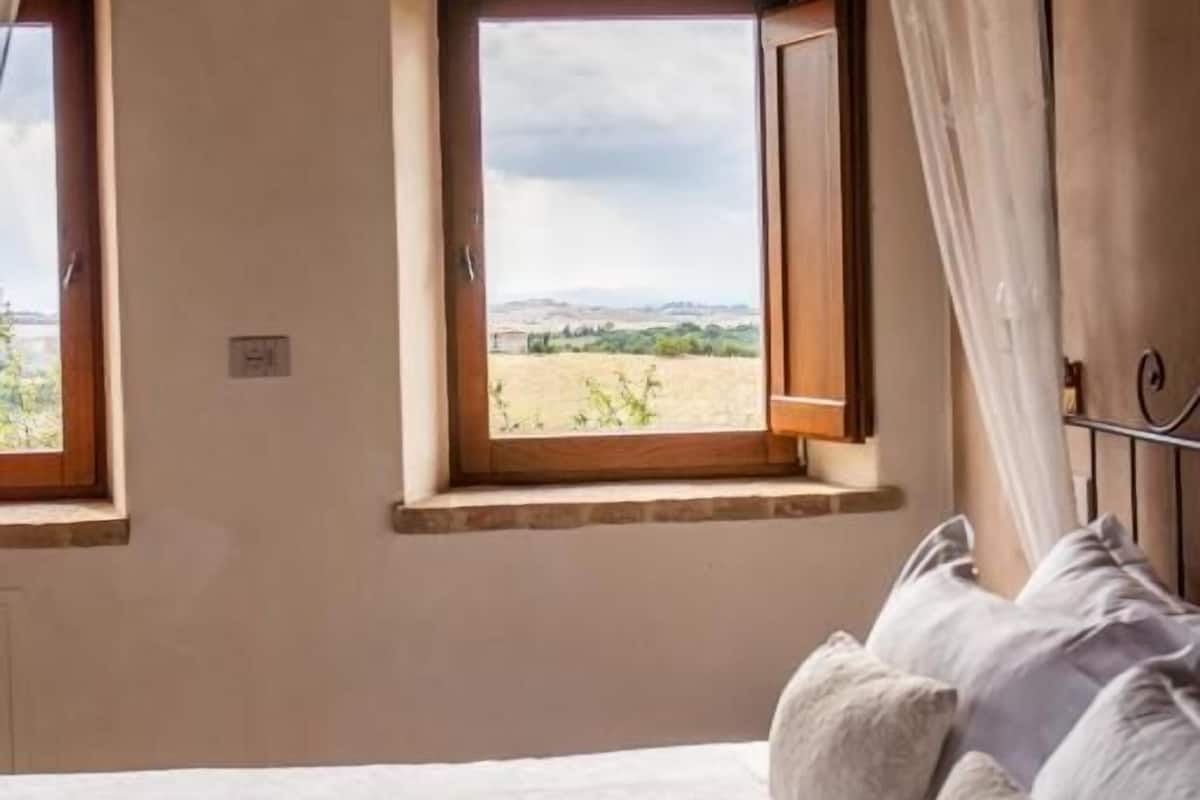 A bedroom with a bed, two windows, and a view of the countryside.