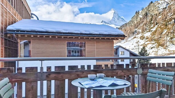Ein Holzbalkon mit Tisch und Stühlen, mit Blick auf einen Berg und ein Gebäude mit verschneitem Dach.