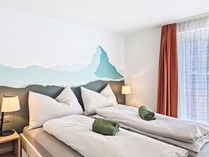 A hotel room with a bed, two pillows, a nightstand with a lamp, a wall with a mountain design, and a window with curtains. - Nice apartment for 4 guests with WIFI, TV and balcony (Zermatt)