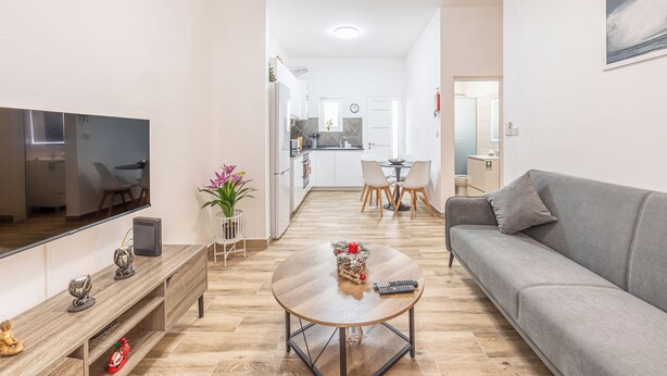 Una sala de estar moderna con un televisor de pantalla plana, un sofá gris, una mesa de centro y una zona de comedor con sillas y una mesita.