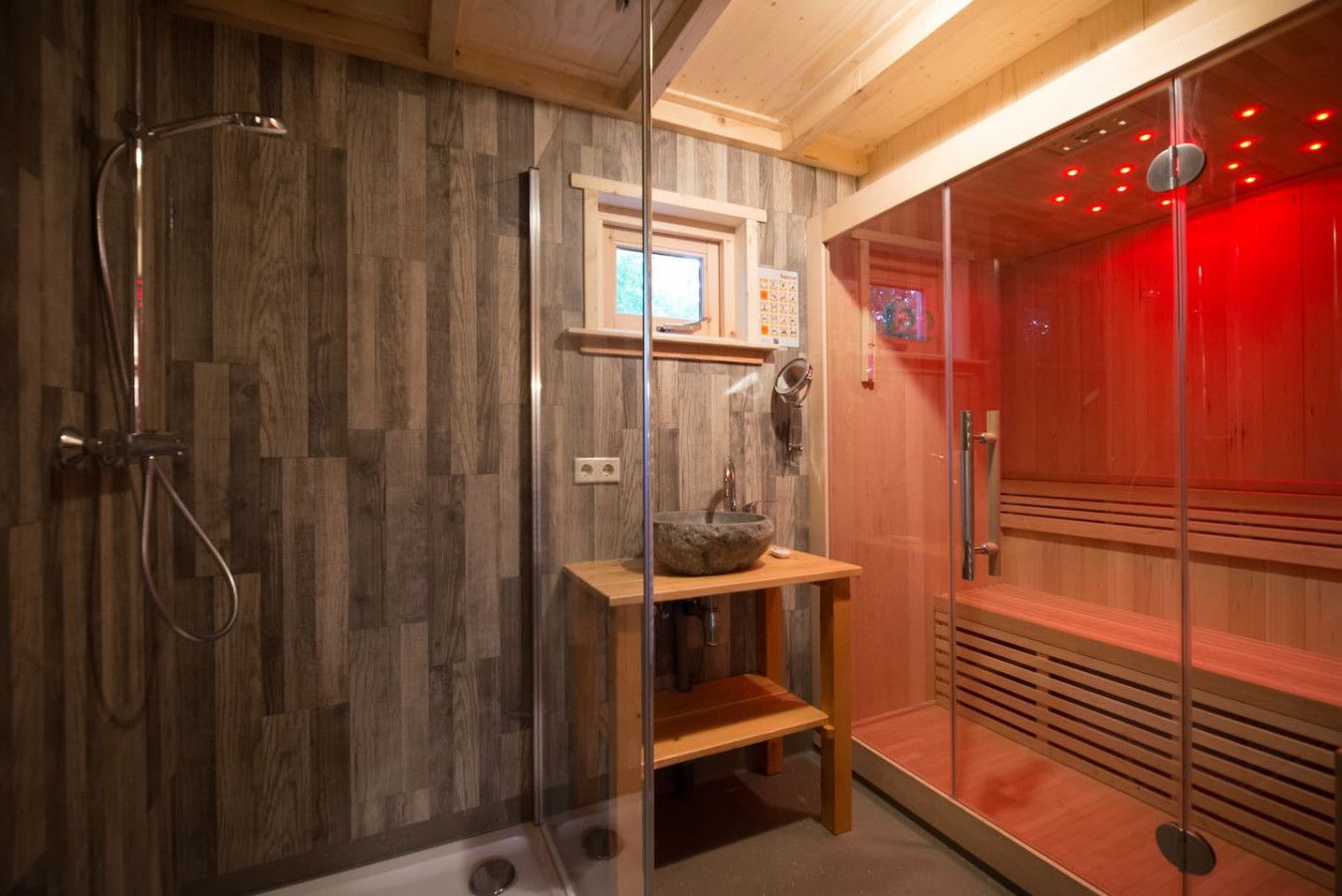 Een moderne badkamer met een houten wand, een glazen douchecabine, een houten wastafel met een stenen wasbak en een sauna met rode verlichting.