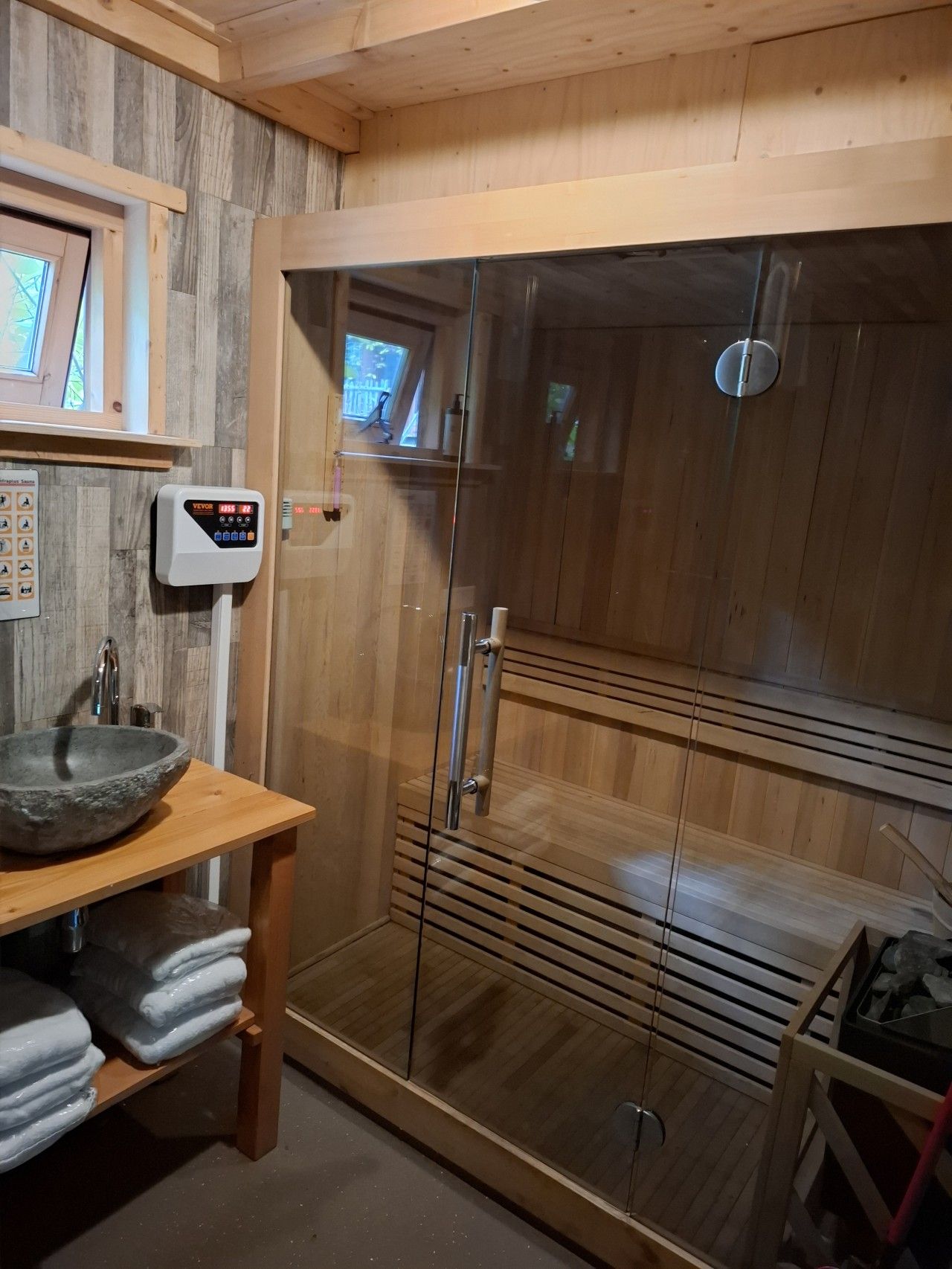 Un sauna avec un intérieur en bois, une porte en verre, un panneau de commande et un lavabo avec des serviettes.