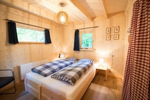 A wooden cabin bedroom with a bed, two bedside tables, a radiator, and a window with curtains.