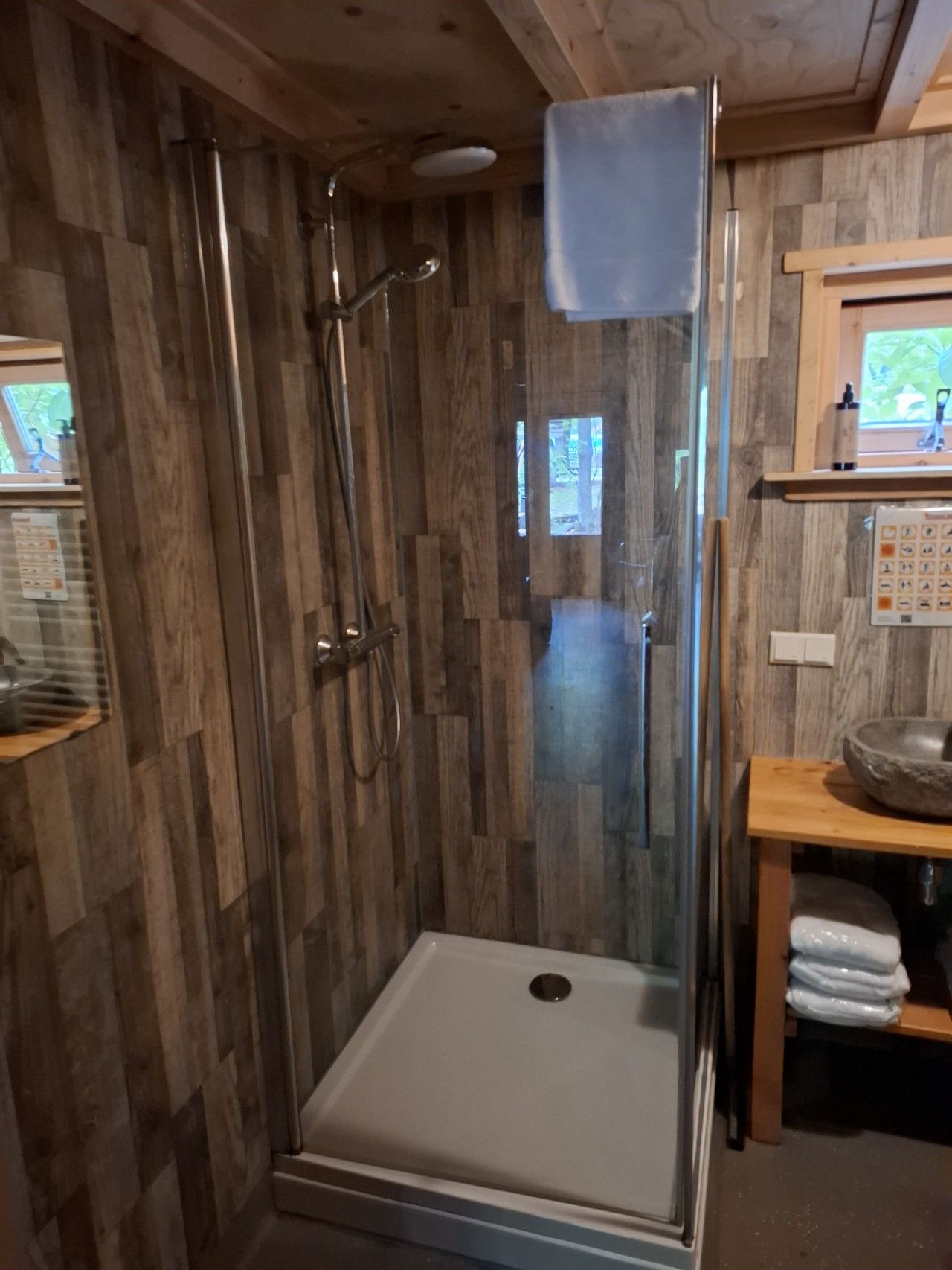 A glass-enclosed shower with a white floor, wooden walls, and a wooden shelf with towels.