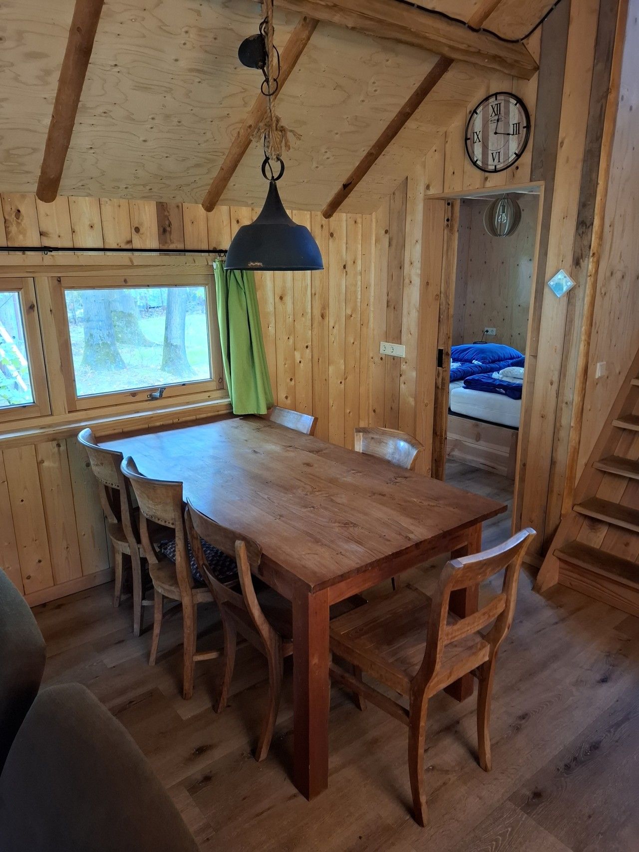 Een houten interieur met een eettafel, stoelen, een hanglamp, een klok en een raam met gordijnen.