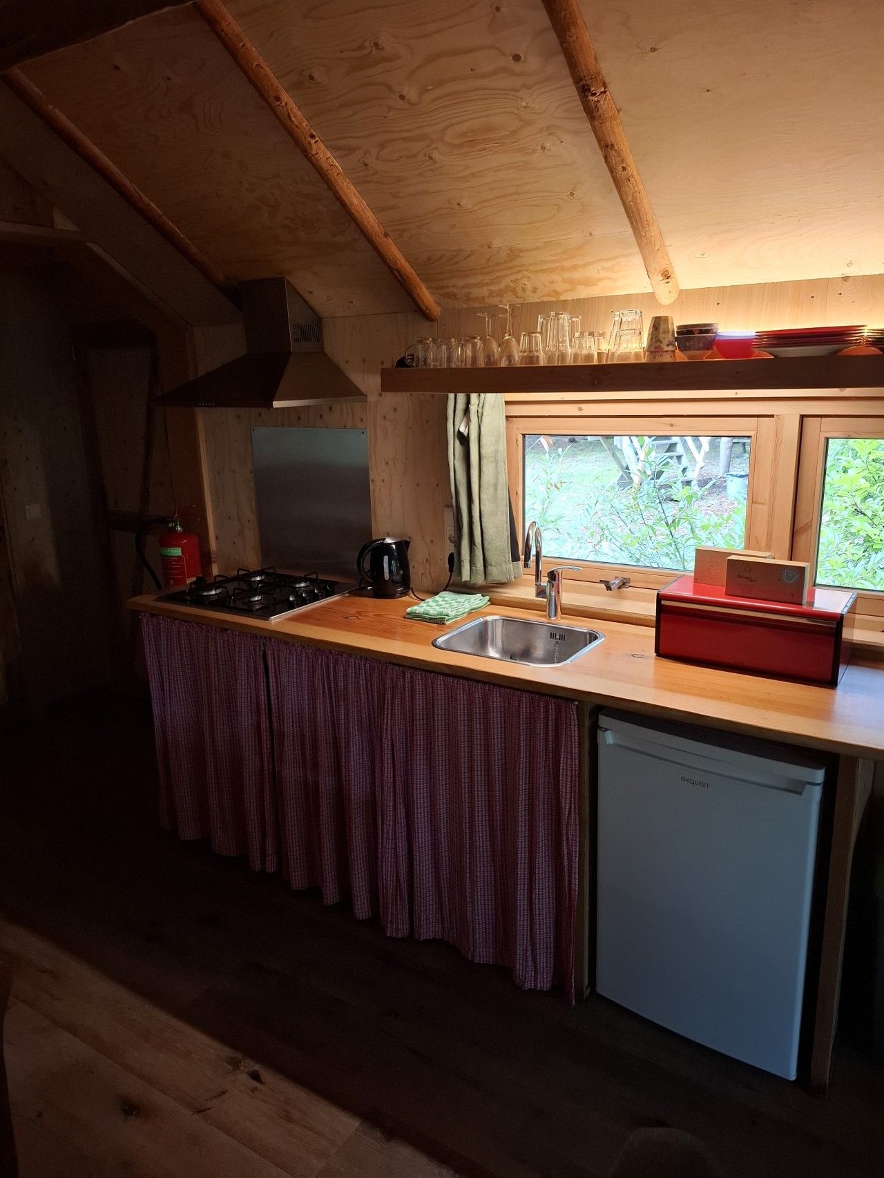 Une cuisine compacte dotée d’un plan de travail en bois, d’un évier, d’un lave-vaisselle et d’une fenêtre donnant sur un espace verdoyant.