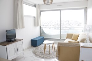 A modern living room with a large window, a sofa, a coffee table, a TV on a cabinet, and a blue bench. - Studio with harbour view, close to the beach - Carnon ideal for short stays (Mauguio)
