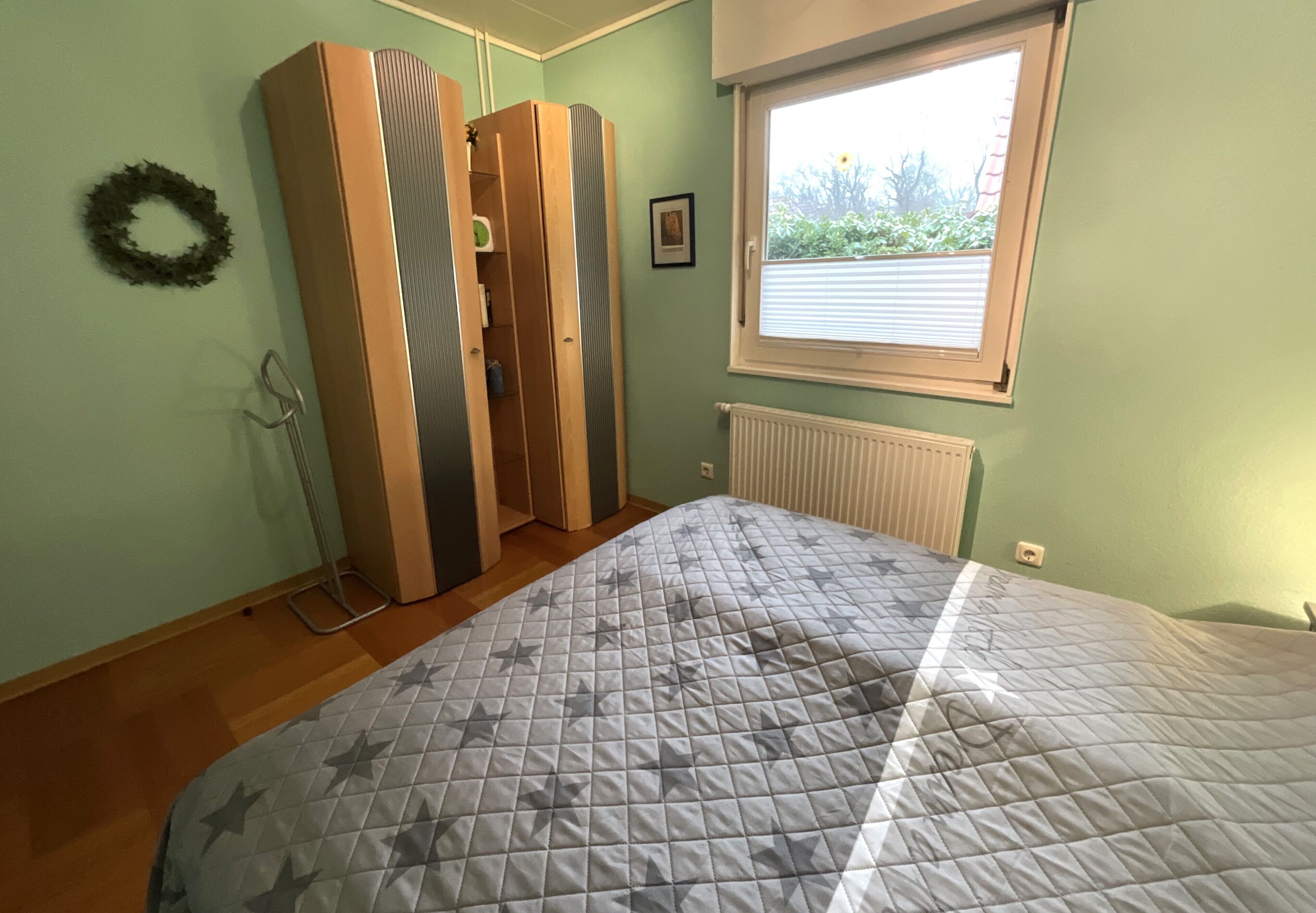 Ein Schlafzimmer mit Bett, Heizkörper, Fenster mit Jalousien, Kleiderschrank und Wandkranz.