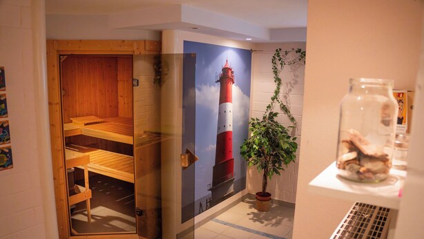 Sala de sauna com bancos de madeira e porta de vidro, um cartaz de um farol e uma planta em vaso.
