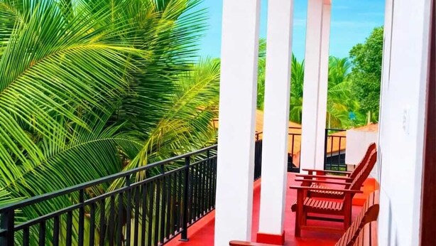A covered outdoor seating area with wooden benches and a red floor, surrounded by a black railing and palm trees. - Wilpattu Gagana Guest  (Pahala Maragahawewa)
