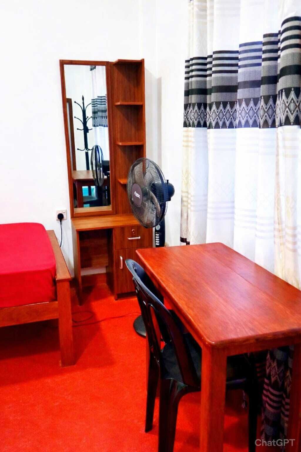 A compact room with a wooden desk, a red bed, a mirror, and a standing fan.