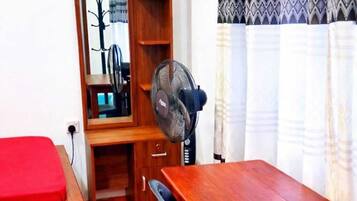 A compact room with a wooden desk, a red bed, a mirror, and a standing fan.