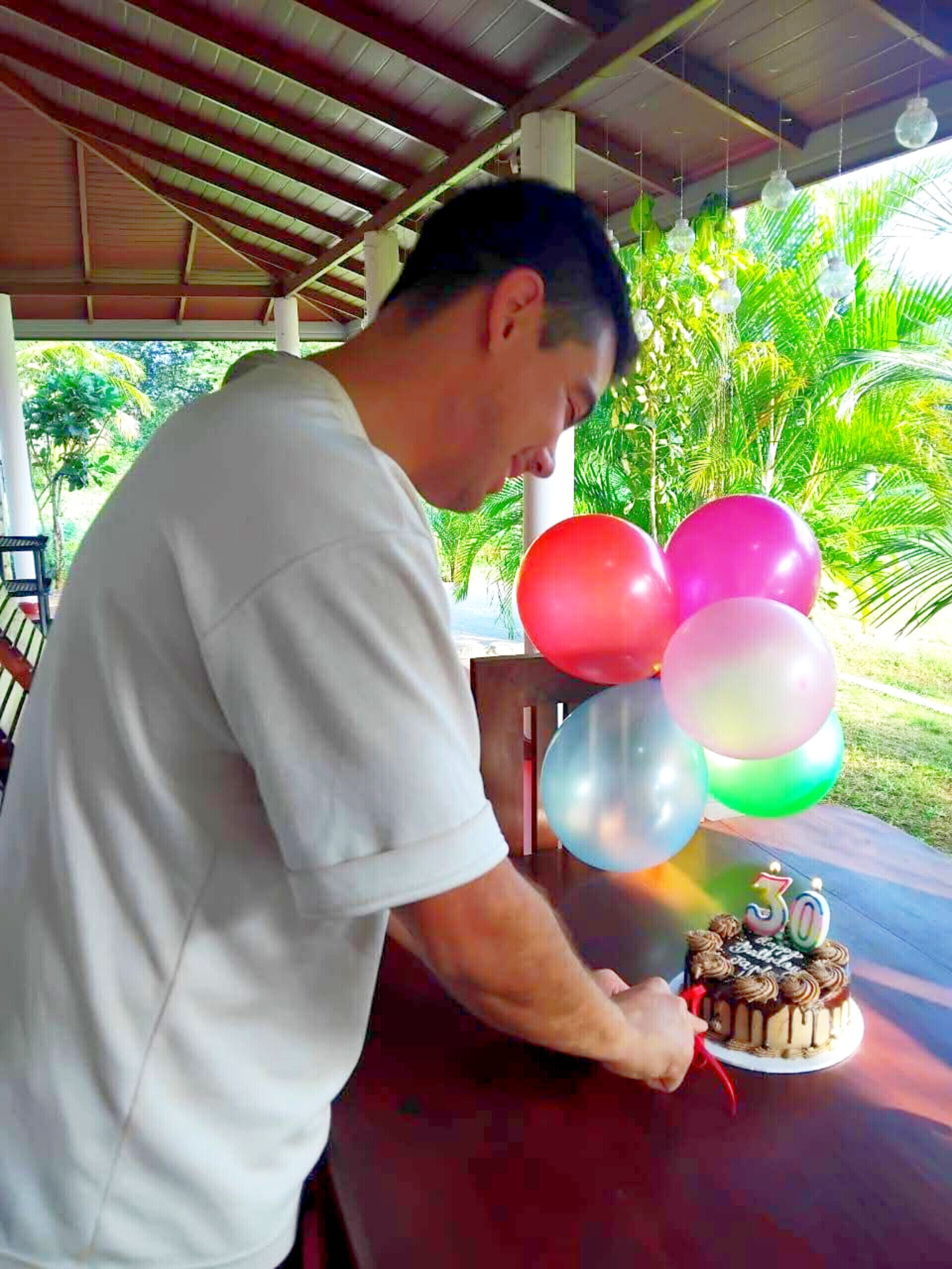 Um homem corta um bolo de aniversário com uma vela em formato de '30' sobre uma mesa, sob um espaço coberto, com vista para áreas verdes.