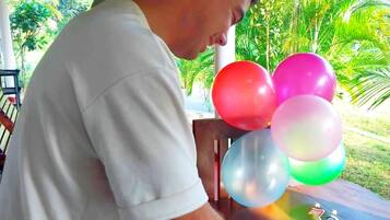 Um homem corta um bolo de aniversário com uma vela em formato de '30' sobre uma mesa, sob um espaço coberto, com vista para áreas verdes.