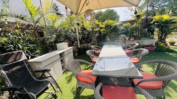 An outdoor patio with a long table and wicker chairs, surrounded by potted plants and a pergola.