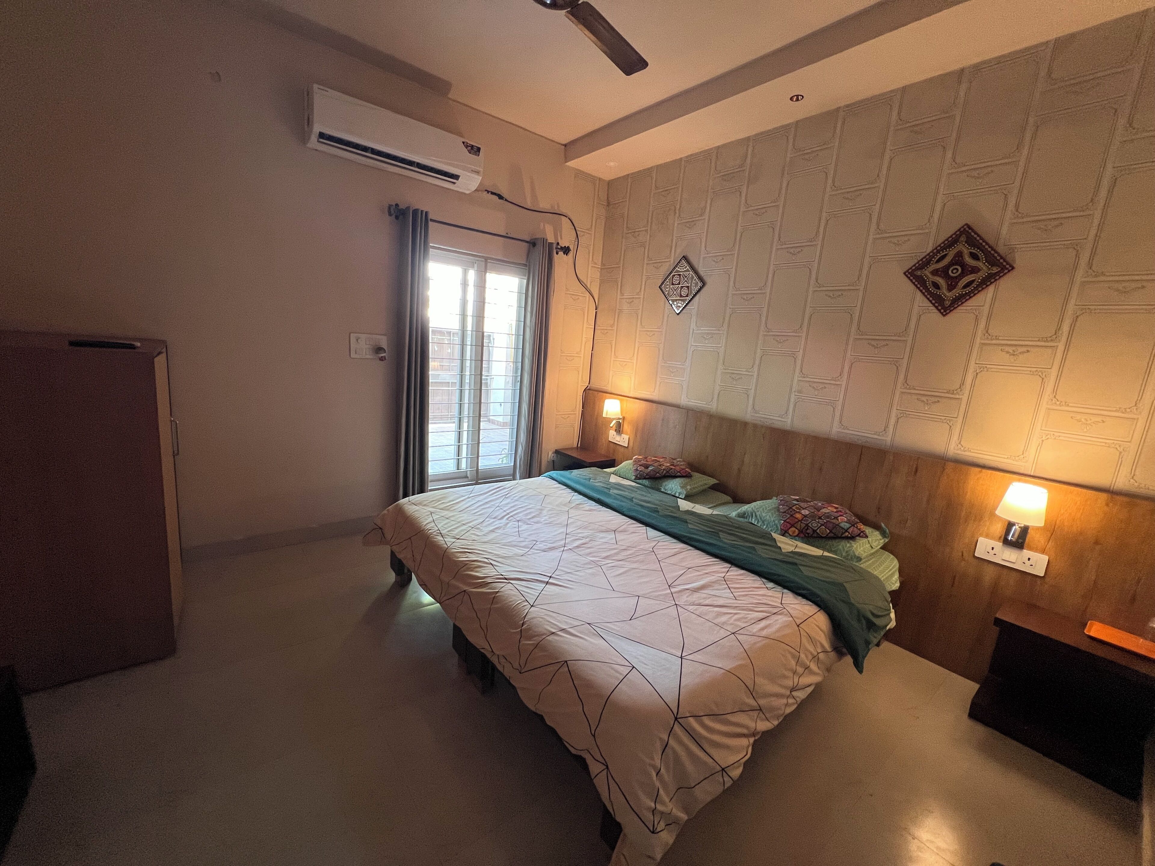 Quarto com cama, mesa de cabeceira, luminária na parede, ar condicionado e janela com cortinas.
