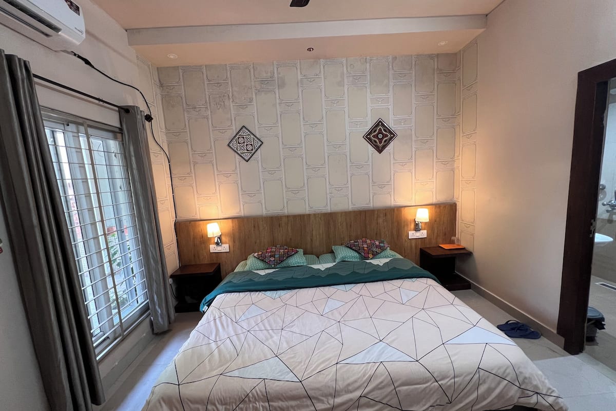 A bedroom with a bed, two bedside lamps, a window with curtains, and patterned wall decor.