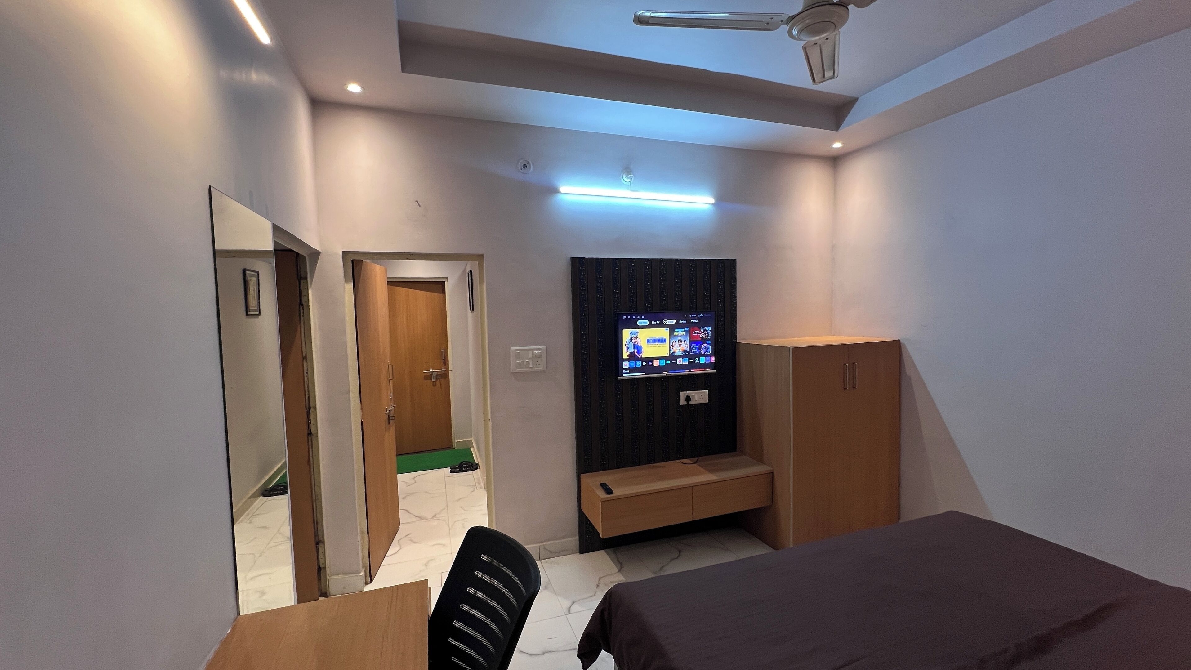A modern bedroom with a bed, a desk, a TV mounted on the wall, and a ceiling fan.