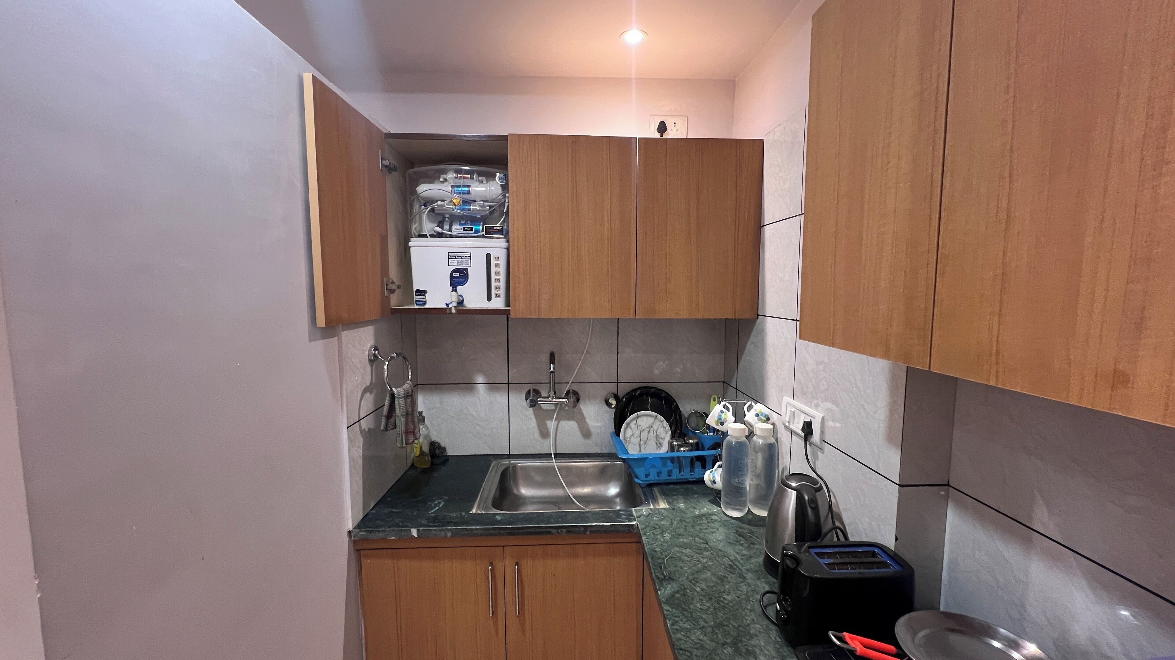 A compact kitchen with wooden cabinets, a stainless steel sink, a kettle, a toaster, and a blue dish rack.