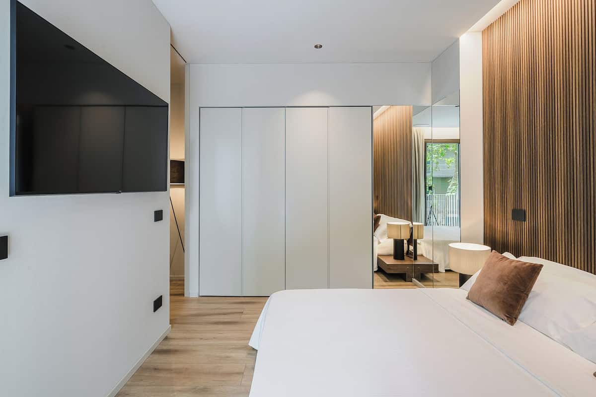 A modern bedroom with a large bed, a TV mounted on the wall, a wardrobe, and a mirror reflecting the room.