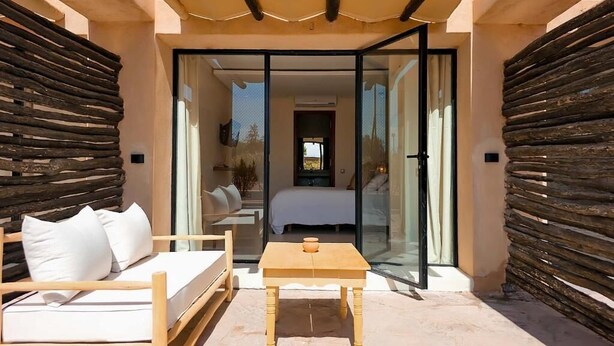 A modern outdoor seating area with a wooden table and sofa, a glass door leading to a bedroom, and a wooden fence. - Villa Katia (Marrakech)