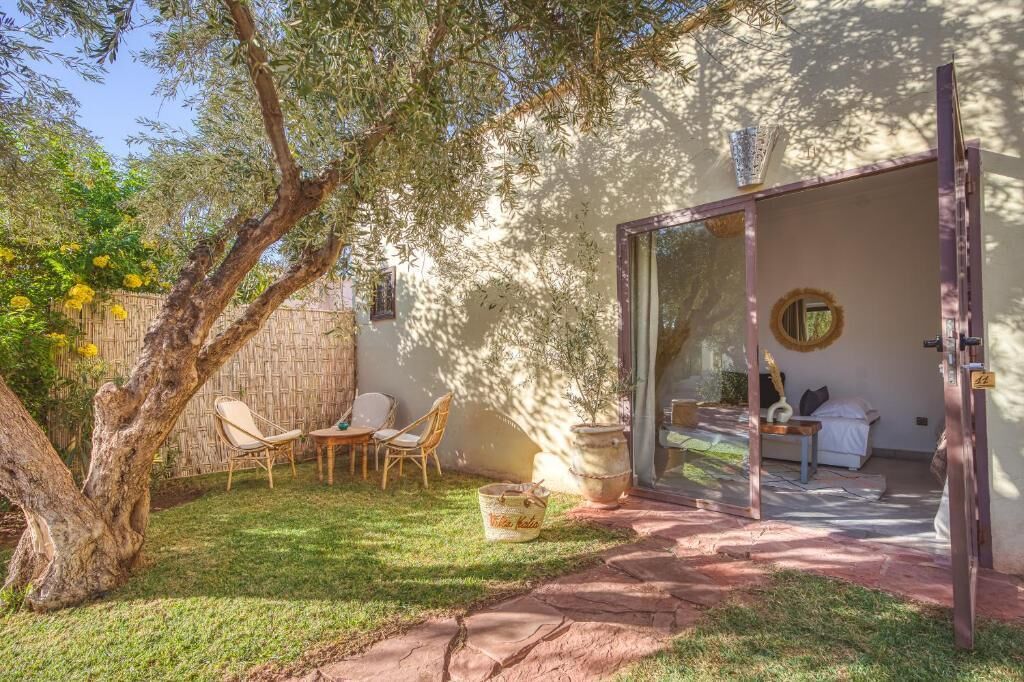 A cozy outdoor seating area with a table and chairs, a large potted plant, and a view into a room with a bed and a round mirror.