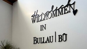 Ein Willkommensschild mit dem Schriftzug "Willkommen in Bullau | BÜ" an einer weißen Wand, ein kleines haussymbolisches Schild mit Text sowie ein Regal mit Pflanze und Stofftier.
