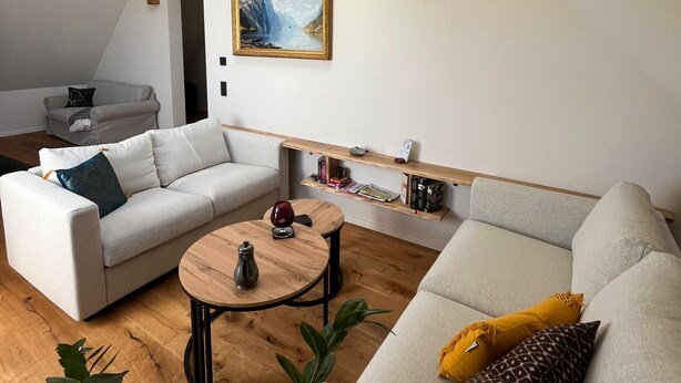 Ein modernes Wohnzimmer mit hoher Decke, Holzbalken, einem Gemälde, einem Sofa, einem Couchtisch und einem Bücherregal.
