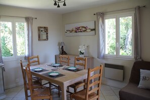 A dining room with a wooden table set for four, surrounded by chairs. There is a window with curtains, a radiator, and a wall-mounted shelf with decorative items. - Gite Casteddu 2 with heated pool for lovers of peace and nature (San Gavino di Carbini)
