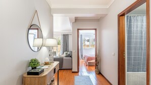 Ein Flur mit Holzboden, einem Konsolentisch, einer Lampe, einem Spiegel und Blick in ein Schlafzimmer mit Bett und Fenster.