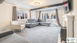 A modern living room with a grey sectional sofa, a white armchair, and a grey carpet. There are large windows with curtains, a chandelier, and a flat-screen TV mounted on the wall. - Cheadle Cottage - Hot Tub and Pool Table! (Alton Towers & Staffordshire Villages)