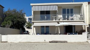 A two-story beachfront building with a balcony, a covered outdoor seating area, and a sandy beach in the foreground. - Dream house ground floor on the beach: prime location at the Mediterranean Sea (Palavas-les-Flots)