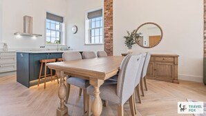 A modern kitchen with a central island, a dining area with a wooden table and chairs, and a brick accent wall. - The Old Chapel - Lower Peaks (Alton Towers & Staffordshire Villages)