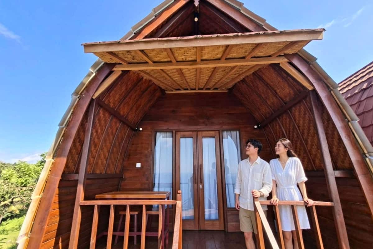 A wooden cabin with a balcony, a wooden table, and a view of the surrounding greenery.