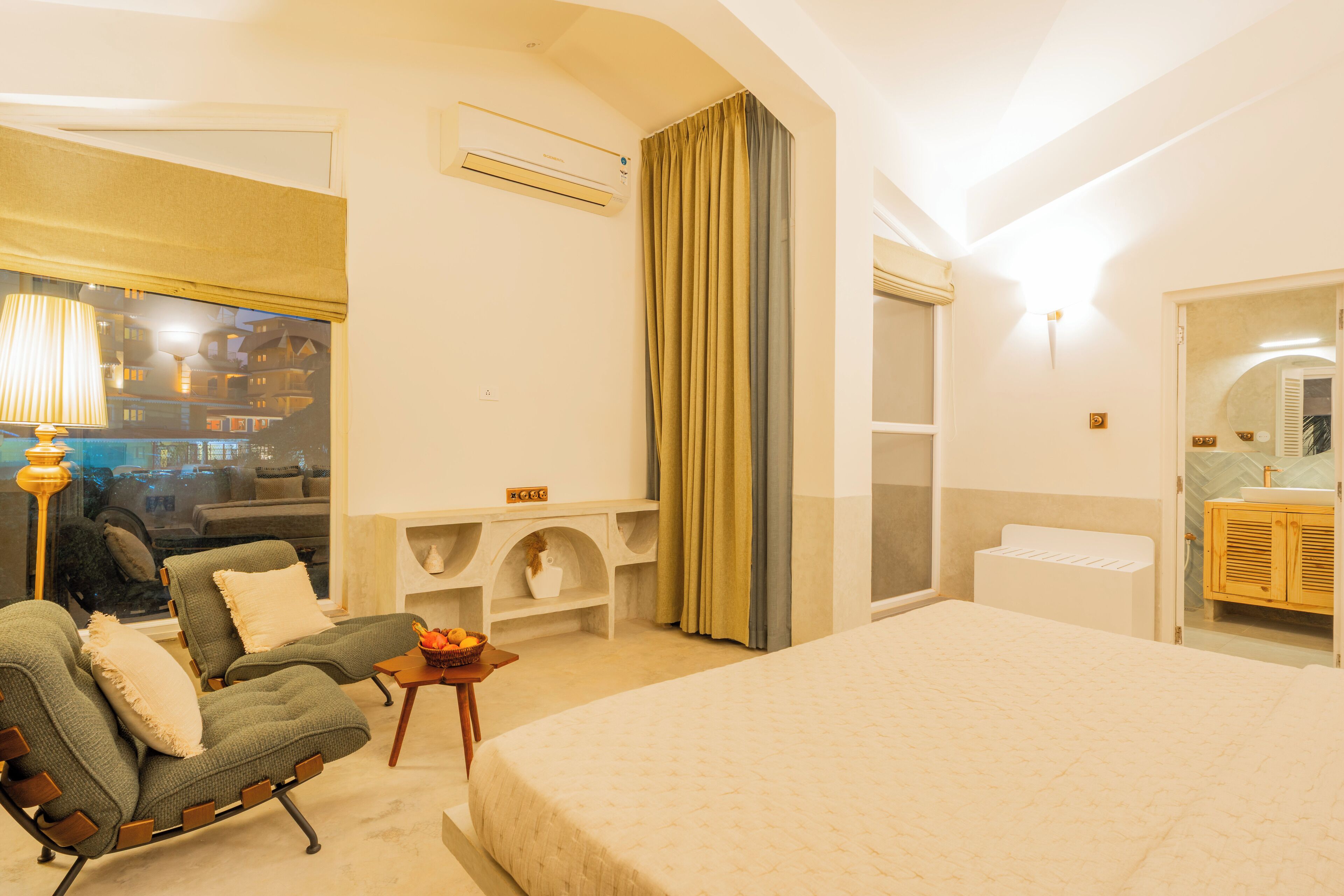 A hotel room with a bed, two armchairs, a small table with a fruit bowl, an air conditioning unit, a mirror, and a view of the outside.