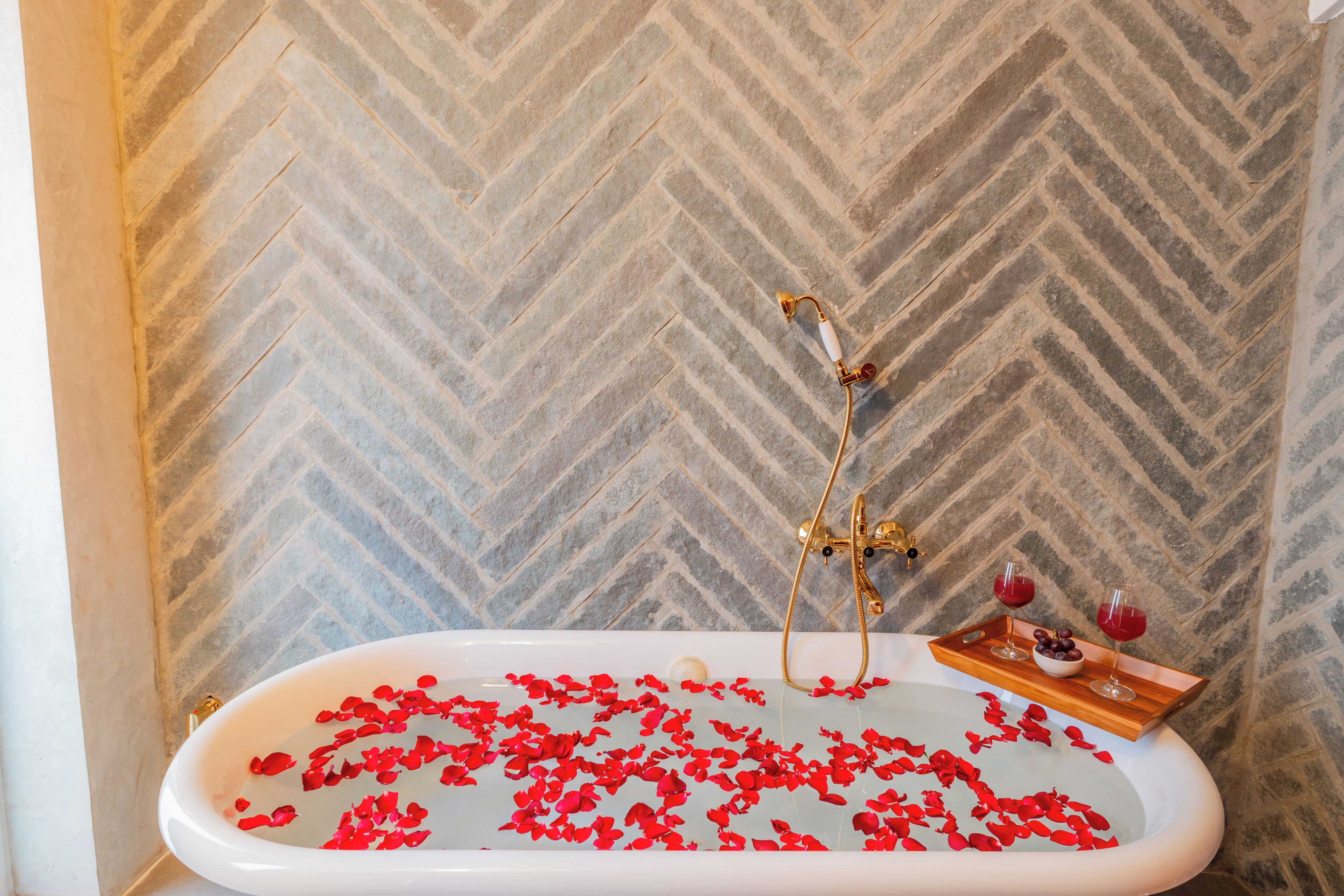 A bathtub filled with rose petals and two glasses of red wine on a tray.