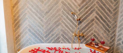 A bathtub filled with rose petals and two glasses of red wine on a tray.