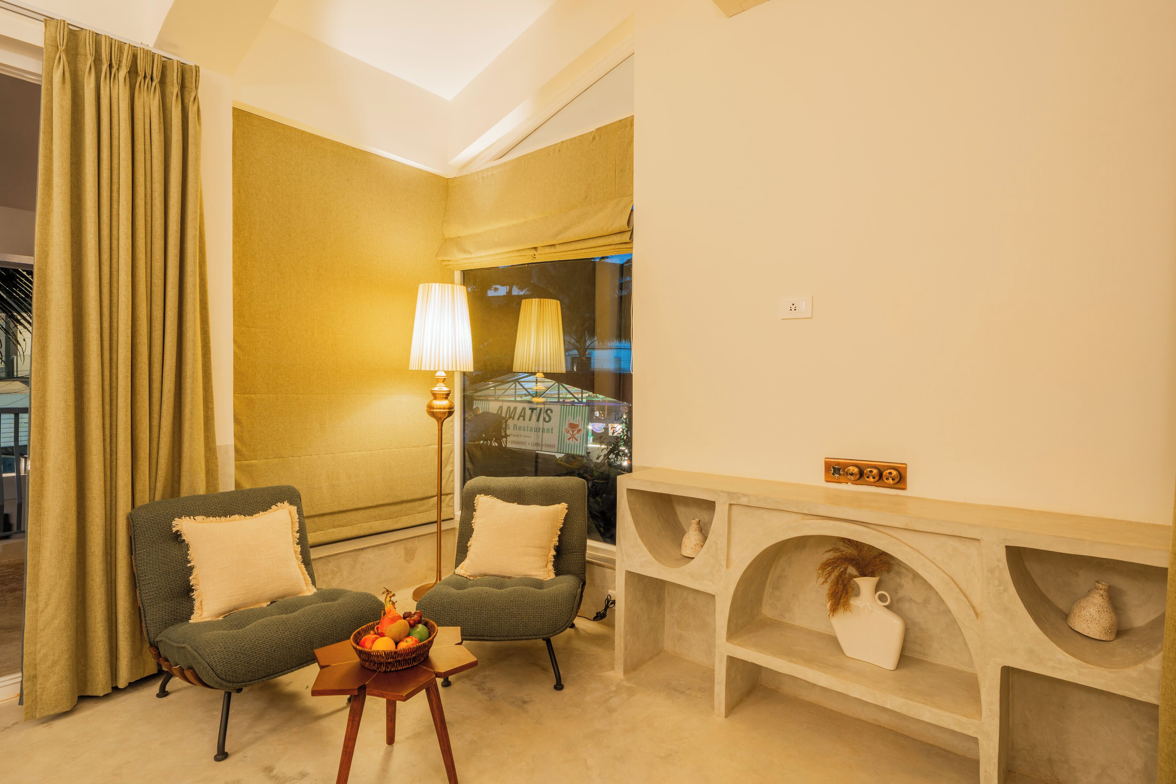 A hotel room with two armchairs, a small table with fruit, a lamp, a mirror, and a view of a building outside.