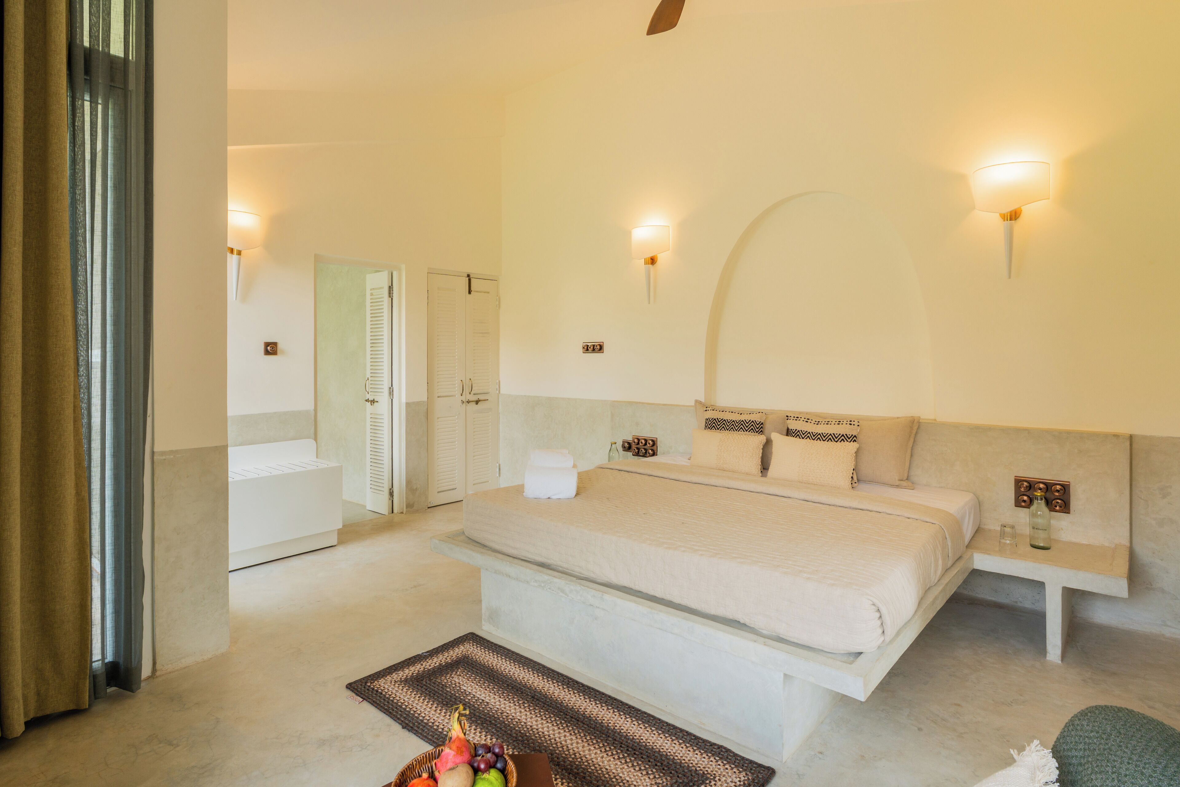 A spacious bedroom with a large bed, a bedside table, and a nightstand. There is a bathroom with a bathtub and a shower visible in the background.
