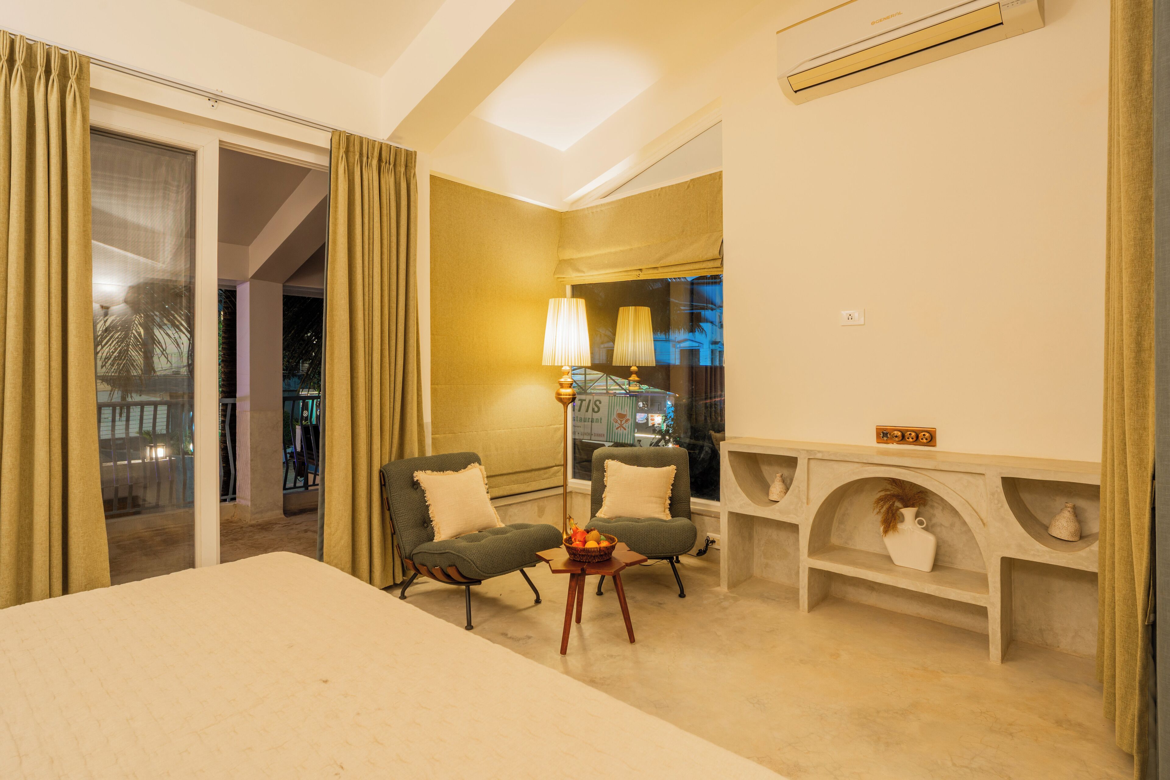 A hotel room with a bed, two armchairs, a small table with a fruit bowl, a lamp, a nightstand, and a view of the city at night.