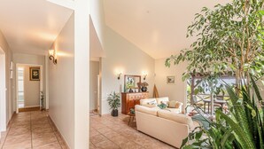 Ein geräumiger Wohnbereich mit einem beigen Sofa, einem Holzschrank und einem Esstisch mit Stühlen. In der Ecke befinden sich Kübelpflanzen und ein großer Baum.